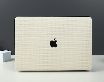 May include: A cream-colored laptop with a textured surface and a black Apple logo. The laptop is on a white surface with a dark gray background. The laptop's surface has a woven pattern.