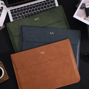 Puede incluir: Tres fundas de cuero para portátiles en verde, azul marino y marrón, cada una con iniciales doradas. También se ven un portátil, una cámara y una taza de café. Las fundas están dispuestas sobre una superficie negra.