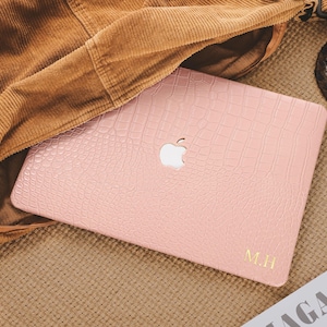 以下が含まれることがあります： クロコダイル柄のピンク色のノートパソコンケース。Appleのロゴと金色のイニシャル「M.H.」が特徴です。ケースは、茶色のコーデュロイジャケットの隣のテクスチャ加工された表面に置かれています。