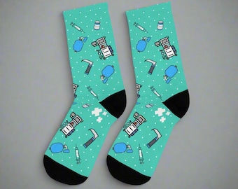 Könnte beinhalten: Ein Paar blaugrüne Socken mit einem Muster aus medizinischen Geräten, darunter eine Spritze, ein Fläschchen, ein Beatmungsbeutel, ein medizinisches Gerät und ein Skalpell. Die Socken haben schwarze Zehen und Fersen.
