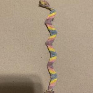May include: A multi-colored woven friendship bracelet with a pink flower charm. The bracelet is made with pink, yellow, and blue threads.