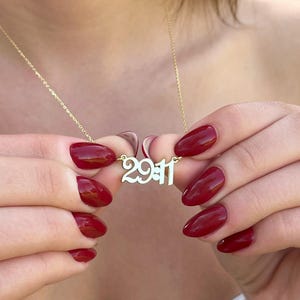 May include: A gold-colored necklace with the numbers "29:11" as a pendant. The necklace is held by a person with red nail polish. The background is blurred, suggesting an outdoor setting.