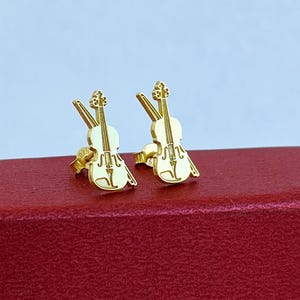 May include: A pair of gold-colored violin-shaped stud earrings. The earrings are displayed on a red surface against a light blue background. The violin studs have detailed features, including the strings and scroll.
