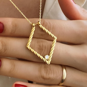 Peut inclure: Un collier doré avec un pendentif en forme de losange. Le pendentif présente un motif torsadé et une petite pierre précieuse transparente. Le collier est porté par une personne portant une chemise blanche. Le design du pendentif est complexe.