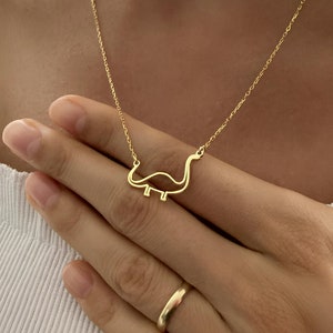 Könnte beinhalten: Eine goldene Kettenkette mit einem zarten Dinosaurier-Anhänger. Der Dinosaurier ist in Gold umrandet und hat einen langen Hals und Schwanz.