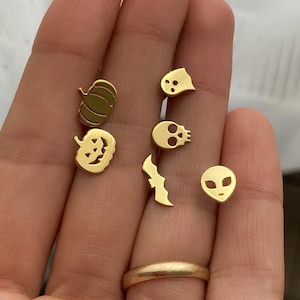 May include: A set of six gold-toned Halloween-themed earrings. The earrings feature a pumpkin, a jack-o'-lantern, a skull, a bat, a ghost, and an alien.