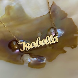 May include: A gold necklace with the name "Isabella" spelled out in cursive script. The necklace is on a white and iridescent shell background.
