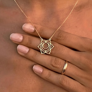 Może przedstawiać: Złoty naszyjnik z delikatnym wisiorem w kształcie stylizowanego kwiatu lotosu. Wisiorek jest symbolem czakry sakralnej, która jest związana z kreatywnością, emocjami i relacjami.