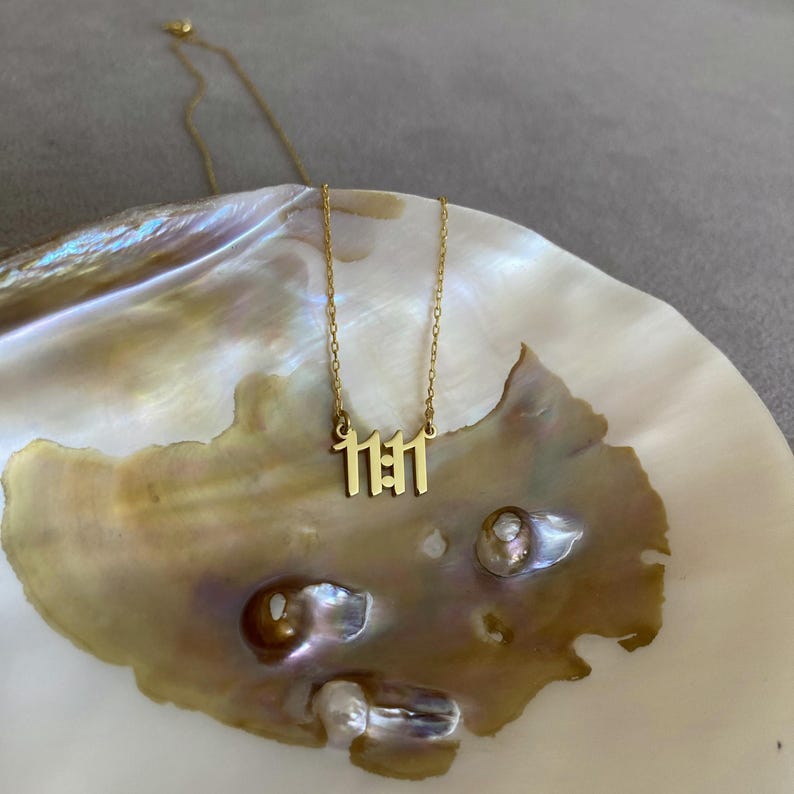 K&ouml;nnte beinhalten: Eine goldene Halskette mit einem Anh&auml;nger, auf dem "1111" steht. Die Halskette liegt auf einer wei&szlig;en und goldenen Muschel.