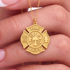 May include: A gold-toned pendant necklace featuring a circular Maltese cross emblem. The center displays a fire helmet, axe, and ladder. The top banner reads FIRE and the bottom banner reads DEPT. A ladder and a fire hydrant are on the sides.