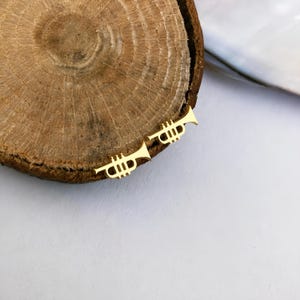May include: A pair of gold-colored trumpet-shaped earrings. The earrings are small and detailed, with a classic trumpet design. They are placed on a wooden surface with a light-colored background.