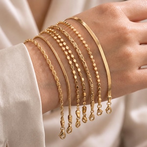 May include: A collection of six gold-toned bracelets featuring various chain styles, including a rope chain, a flat herringbone chain, a beaded chain, and a woven link chain. Each bracelet is finished with a small lobster claw clasp closure.