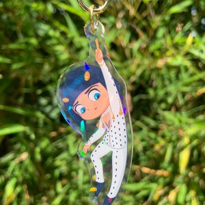 May include: A clear acrylic keychain featuring a cartoon character in a white suit with black polka dots. The character has blue hair and is pointing upwards. The keychain has a gold-colored clasp and a star-shaped charm.