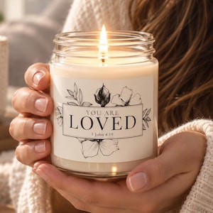 May include: A clear glass jar candle with a lit wick and a white label featuring black floral illustrations and the text "YOU ARE LOVED 1 John 4:19". The candle is filled with off-white wax and held in place by a person wearing a cream knit sweater.