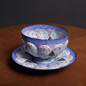 May include: A blue and white ceramic bowl with intricate floral designs sits on a matching saucer. The bowl has a scalloped rim and a raised base.