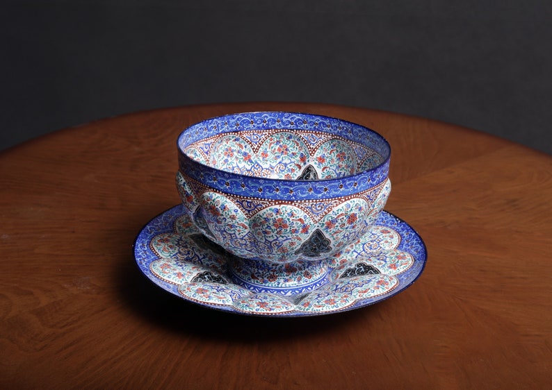 May include: A blue and white ceramic bowl with intricate floral designs sits on a matching saucer. The bowl has a scalloped rim and a raised base.