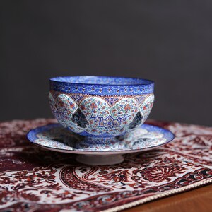 May include: A blue and white ceramic bowl with intricate floral and geometric patterns sits on a matching saucer. The bowl and saucer are on a red and white patterned fabric.