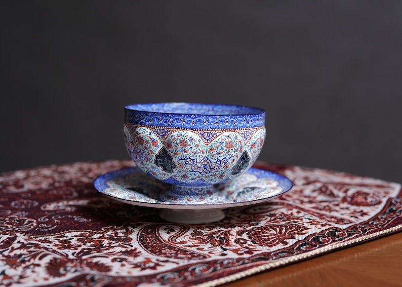 May include: A blue and white ceramic bowl with intricate floral and geometric patterns sits on a matching saucer. The bowl and saucer are on a red and white patterned fabric.