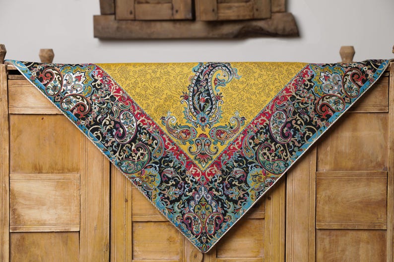 May include: A yellow tablecloth with a paisley pattern border. The tablecloth has a black, red, blue, and green paisley pattern. The tablecloth is on a wooden surface.