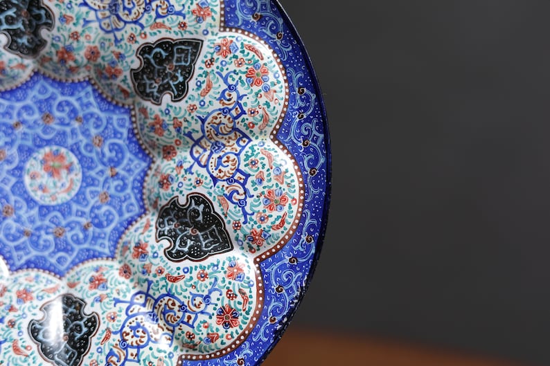 May include: A blue and white ceramic plate with intricate floral and geometric designs. The plate has a scalloped edge and is decorated with black, red, and green accents.