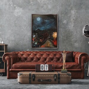 May include: A painting of a dark, moonlit landscape with a large, round moon in the sky. The painting is framed in a silver frame and hangs on a gray wall above a brown leather sofa. The sofa is in a living room setting with a wooden coffee table and a decorative metal stand with a spotlight.