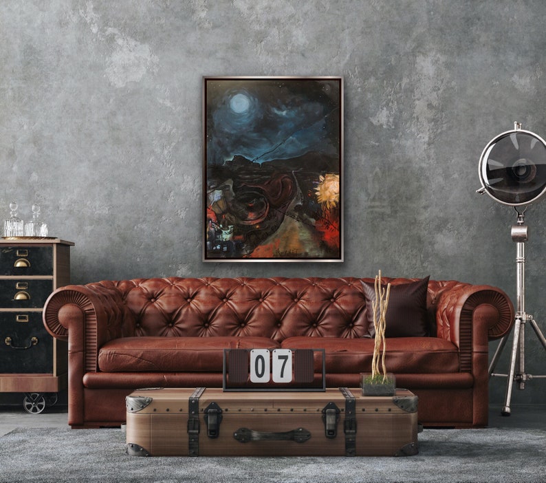 May include: A painting of a dark, moonlit landscape with a large, round moon in the sky. The painting is framed in a silver frame and hangs on a gray wall above a brown leather sofa. The sofa is in a living room setting with a wooden coffee table and a decorative metal stand with a spotlight.