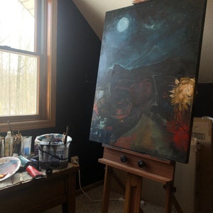 May include: A wooden easel with a canvas painting of a night sky with a moon and stars. The painting is on a wooden frame and is propped up on the easel. There are paint brushes and a bucket of paint on a table in the foreground.