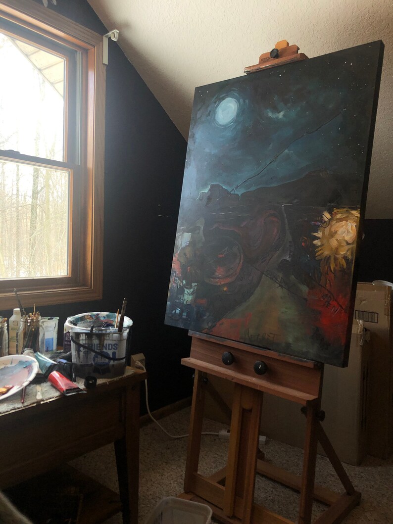 May include: A wooden easel with a canvas painting of a night sky with a moon and stars. The painting is on a wooden frame and is propped up on the easel. There are paint brushes and a bucket of paint on a table in the foreground.