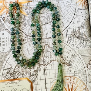 Puede incluir: Un collar mala de ágata verde con un borla verde. El collar está hecho de cuentas redondas y se coloca sobre un mapa vintage.