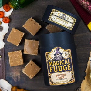 Può includere: Un contenitore cilindrico nero con la scritta "Magical Fudge" e un'illustrazione di un mago, accompagnato da quadrati di fudge. Il coperchio del contenitore ha anche l'etichetta "Magical Fudge". Il fudge è al burro.