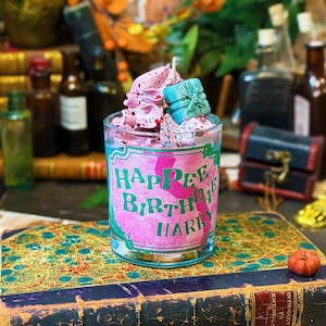 May include: A glass candle with pink and white wax, topped with a small blue gift box. The candle has a label that reads "Happee Birthday Harry" in green and pink. The candle is placed on a decorative book.