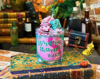 May include: A clear glass candle with pink and white wax, topped with a small blue gift box. The candle has a label that reads "Happee Birthday Harry" in green and pink. The candle sits on a decorative book.