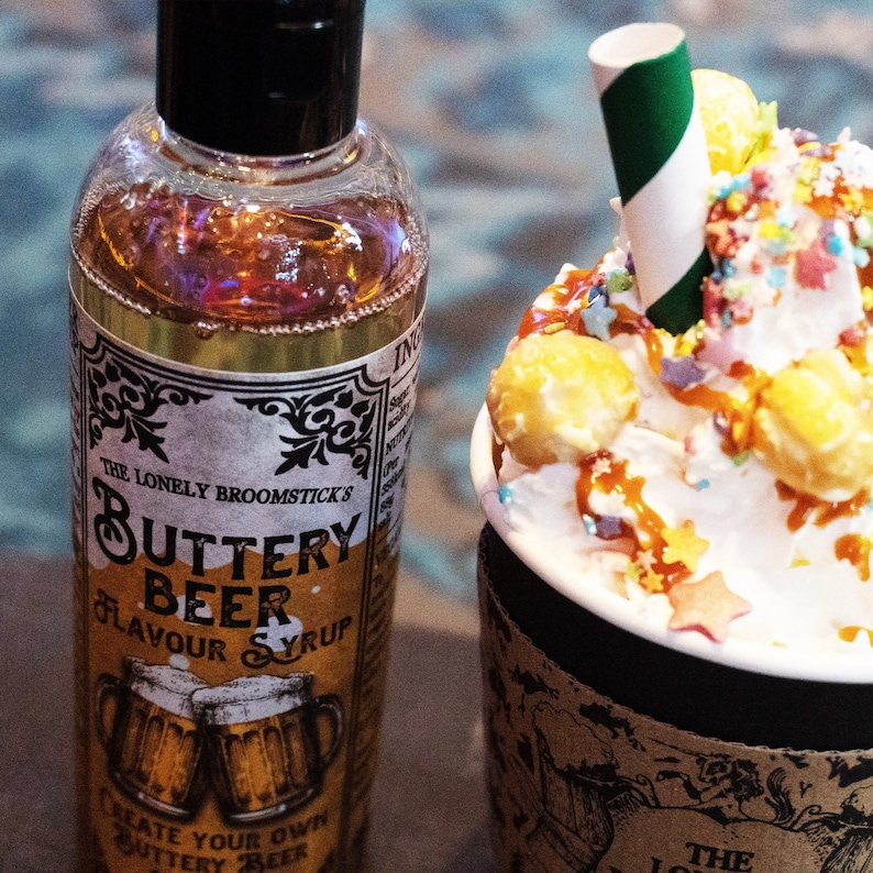 May include: A clear glass bottle with a black cap and a label that reads "Buttery Beer Flavor Syrup" by "The Lonely Broomstick's". The bottle is next to a mug filled with whipped cream, caramel sauce, and popcorn, topped with colorful sprinkles.