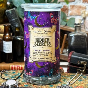 Puede incluir: Un tarro de vela cilíndrico con tapa de cristal, con una etiqueta morada con mariposas, lunas y el texto "Hidden Secrets Witchy Edition". La vela pesa 600g y tiene una duración de 90 horas.