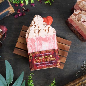 May include: Handmade soap bar with a pink and white swirl pattern, topped with a whipped cream effect and a red heart. The label says "Charming Love Potion". The soap is on a wooden soap dish.