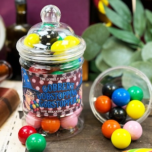 Op de afbeelding: Een glazen pot gevuld met kleurrijke Gobstopper-snoepjes, met een etiket met een illustratie van een tovenaar en de tekst "Gobber's Gobstopping Gobstopper". Een kleine kom met snoep staat in de buurt. De snoepjes zijn verschillend gekleurd.
