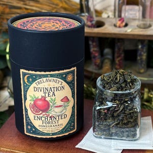 May include: A black cylindrical container of Trelawney's Divination Tea, Enchanted Forest Pomegranate flavor, next to a glass jar filled with loose leaf tea. The container has a decorative label with a pomegranate illustration.