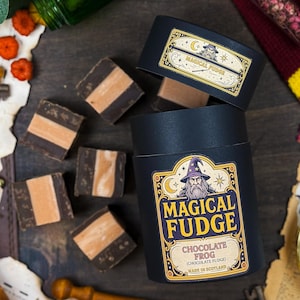 May include: A black container of "Magical Fudge Chocolate Frog" with a wizard illustration, alongside fudge squares with a light brown centre. The container is labelled "Made in Scotland". Fudge pieces are scattered around.