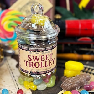 Puede incluir: Un tarro de cristal transparente lleno de coloridas gominolas y chocolates. El tarro tiene una tapa y una etiqueta que dice "Sweet Trolley". Un tren de juguete rojo está en el fondo, junto con una piruleta colorida.