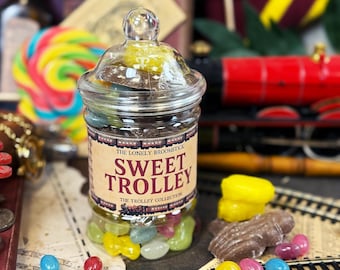 May include: A clear glass jar filled with colorful jelly beans and chocolates. The jar has a lid and a label that reads "Sweet Trolley". A red toy train is in the background, along with a colorful lollipop.