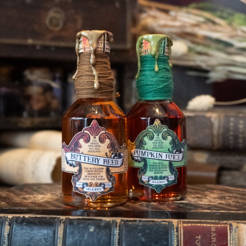 May include: Two glass bottles with gold and green wax seals. The bottles are labeled "Buttery Beer" and "Pumpkin Juice". The labels have a vintage design with ornate lettering and illustrations.