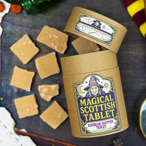 Può includere: Un contenitore cilindrico di cartone con la scritta "Magical Scottish Tablet" e un'illustrazione di strega, insieme a pezzi di fudge marrone chiaro. Il contenitore è aperto, rivelando più fudge e un coperchio con un design simile.