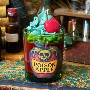 Peut inclure: Un cocktail Poison Apple dans un verre à bord vert. La boisson a une base rouge, surmontée de crème fouettée verte et bleue et d'une pomme rouge. Un autocollant Poison Apple est sur le verre.