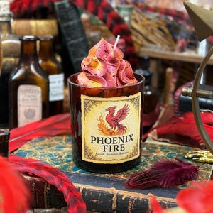 May include: A candle in a dark glass container with a label that reads "Phoenix Fire." The candle has a pink and yellow floral design on top. The label features a red phoenix and flames. The candle sits on a stack of old books.