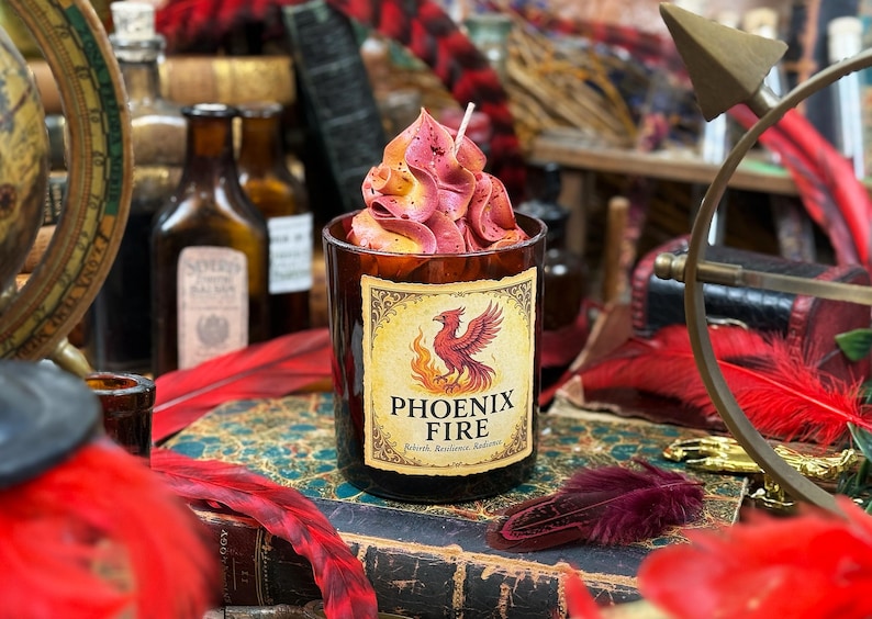 May include: A candle in a dark glass container with a label that reads "Phoenix Fire." The candle has a pink and yellow floral design on top. The label features a red phoenix and flames. The candle sits on a stack of old books.