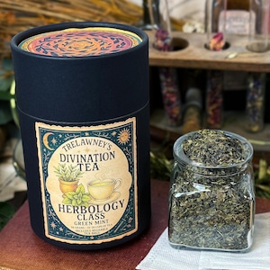 Könnte beinhalten: Ein dunkelblauer zylindrischer Behälter mit Trelawney's Divination Tea, Herbology Class, Green Mint. Ein Glasgefäß mit losem Blatttee steht neben dem Behälter. Der Behälter hat einen dekorativen Deckel.