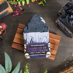 Puede incluir: Jabón escocés hecho a mano con un diseño oscuro y en capas, sobre una jabonera de madera. El jabón está etiquetado como "Draught of Living Death" con gráficos de calaveras. Otros jabones y artículos vintage están en el fondo.