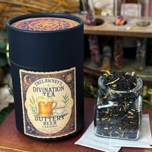 May include: A black cylindrical container of Trelawney's Divination Tea, with a label featuring a beer mug and teacup. A small glass jar of loose tea sits beside it. The tea is labeled "Buttery Beer Caramel". The container has a decorative lid.