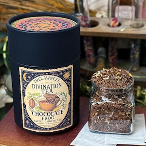 May include: A dark blue cylindrical container of Trelawney's Divination Tea, Chocolate Frog flavor, next to a glass jar filled with loose tea leaves. The label features a frog in a teacup and the text "Sweet Chocolate".