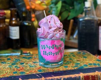 May include: A clear glass candle with a pink and teal label that reads "Happee Birthdae". The candle has a pink, whipped-cream-like top with sprinkles and a small wick. The candle sits on a decorative book.
